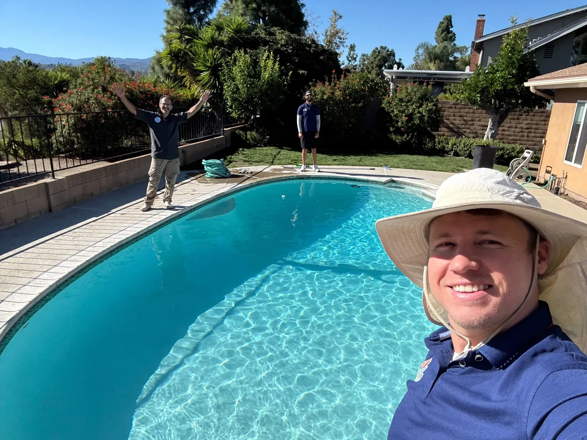 A technician close to the camera with two teammates waving beside a freshly cleaned blue pool