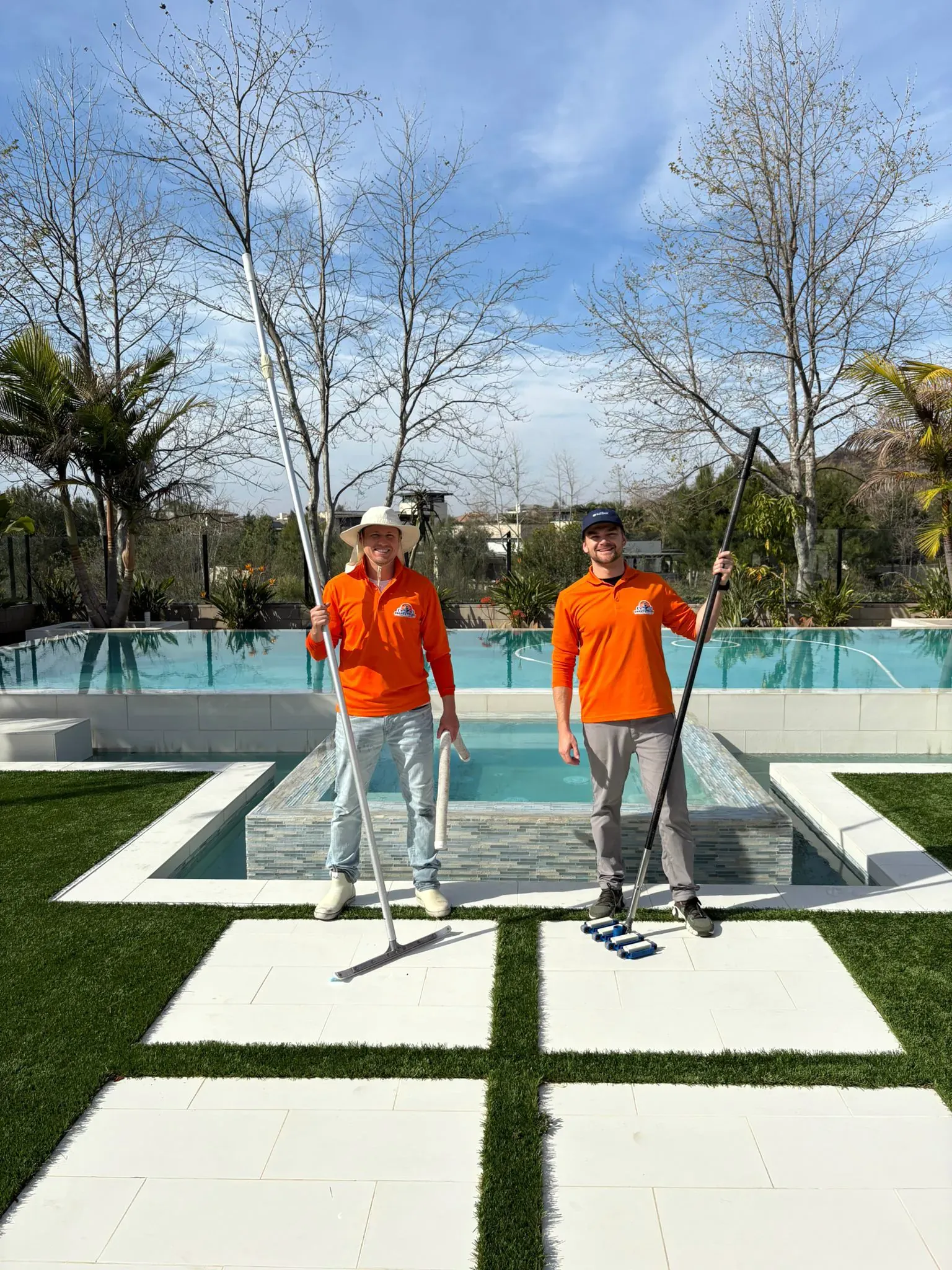 Two technicians holding cleaning poles beside a modern backyard pool and spa