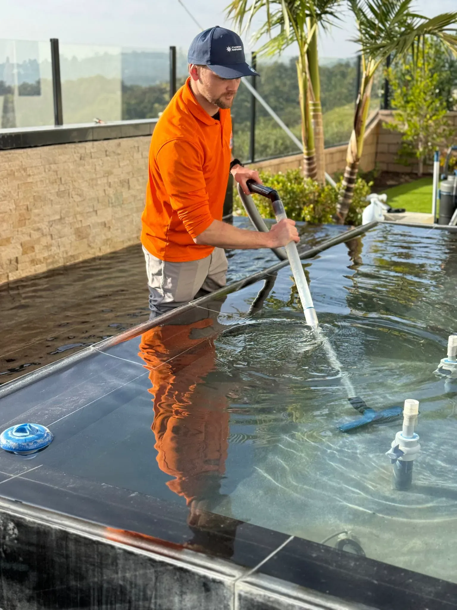 A technician vacuuming the floor of a modern glass-edge infinity pool with a pole