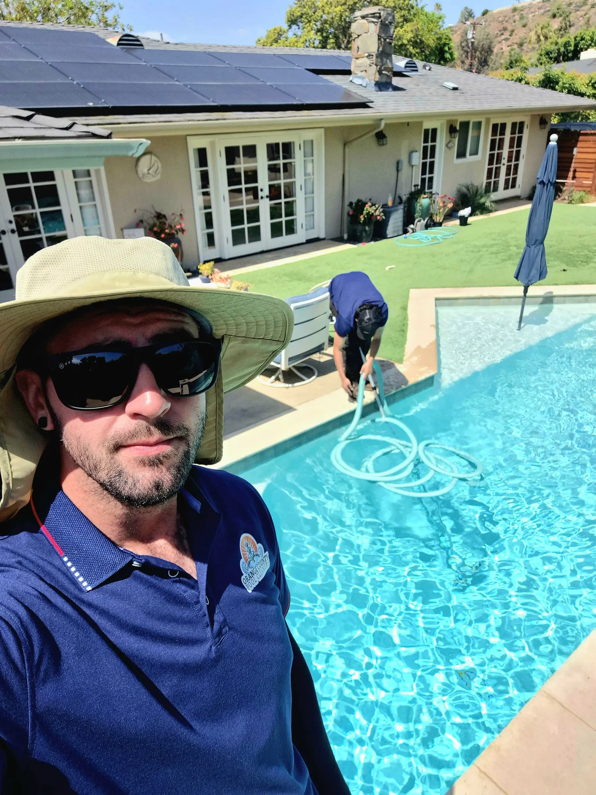 A technician in a bucket hat close to the camera while a teammate vacuums a clear blue pool in the background